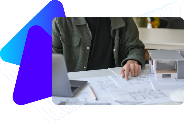 Person in dark jacket sitting at desk with architectural plans and a laptop.