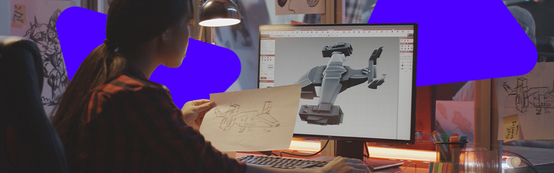 A person with dark hair and a checkered shirt sits at a desk, working on a computer with a large monitor.