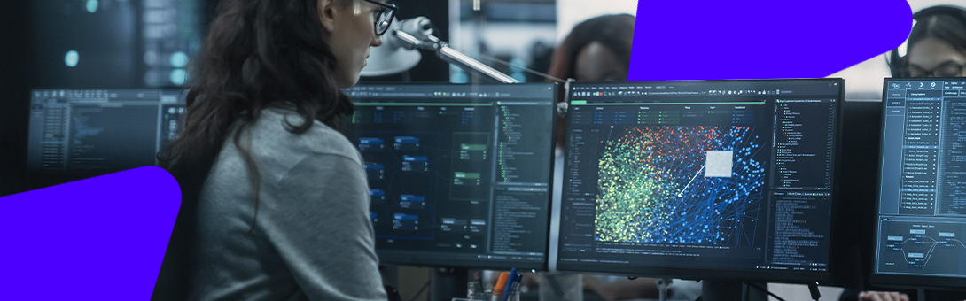 A person with curly hair working at a desk with multiple computer monitors displaying data.
