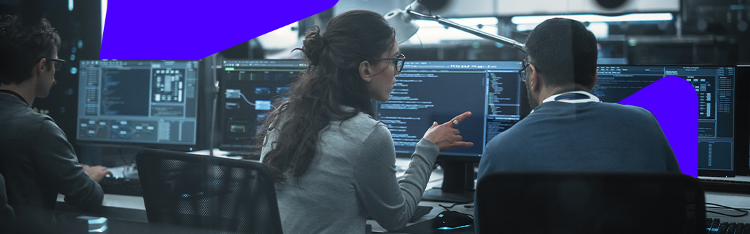Two people working together at a computer in a dimly lit tech workspace.