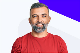 A smiling man with gray hair and beard wearing a red shirt, standing against a white and purple background.