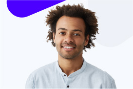 A young man with curly hair and a beard smiling, wearing a light blue shirt against a plain background.