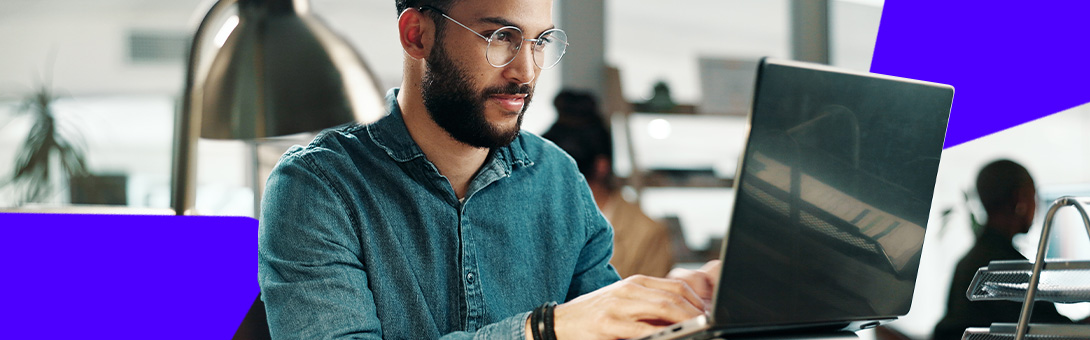 A man with glasses and a beard works on a laptop in a modern office setting.