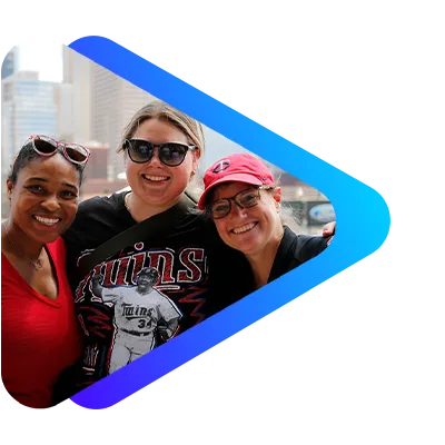 Three smiling women wearing sunglasses and casual clothing outdoors at a sporting event on a sunny day.